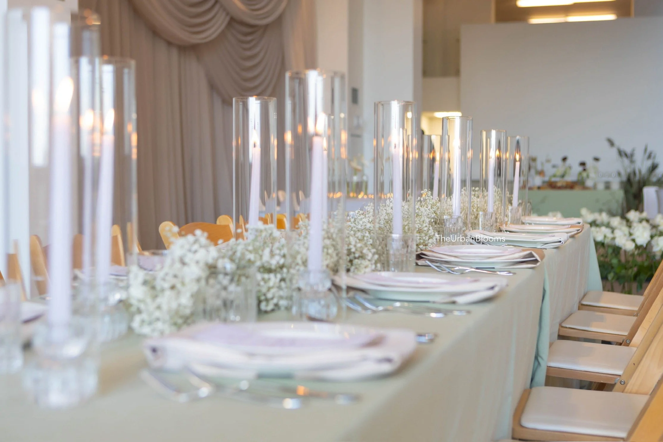 A long dining table set for a special event with candles, white floral arrangements, and tableware in a bright, elegant room with draped curtains